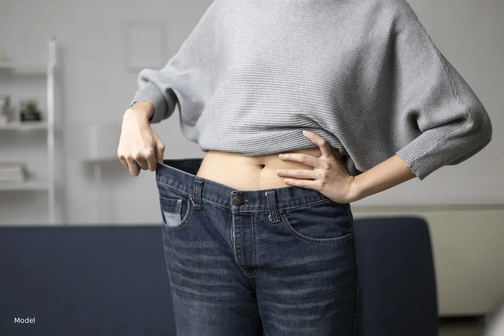 Woman showing slim midsection pulling jeans waistband away, grey sweater